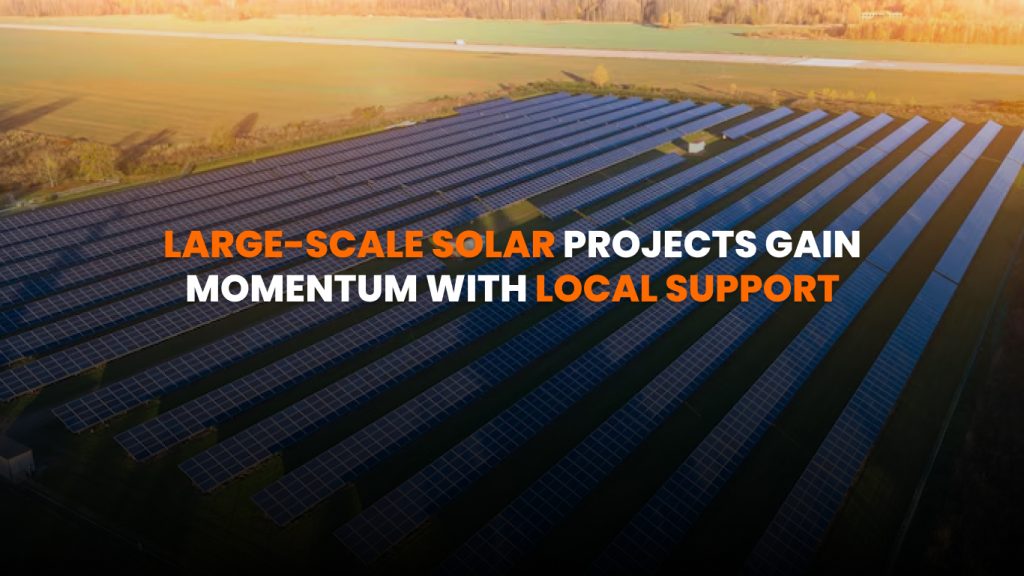 Community solar farm with panels installed on open land under blue sky, with town buildings in the background